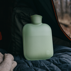 Large green hot water bottle with ribbed design and soft cover, standing on bedding