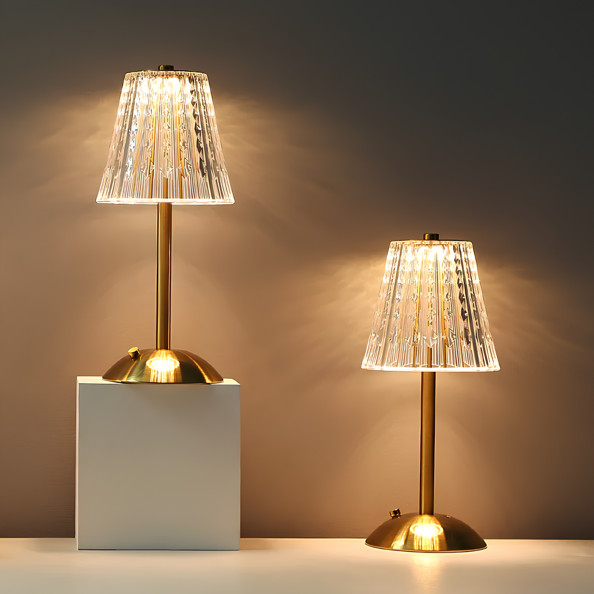 Two gold table lamps with clear pleated crystal shades and round bases, lit on blocks.