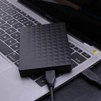 Black external hard drive with geometric face near laptop keyboard, USB plug visible