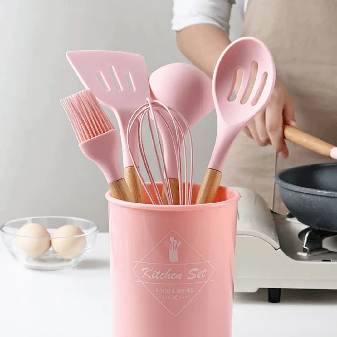 Pink silicone kitchen utensil set with wooden handles in a matching utensil holder cup