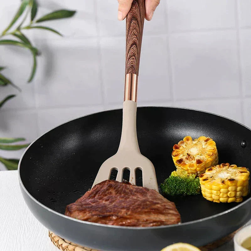 Beige silicone spatula with brown wooden handle flipping meat in a black frying pan