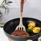 Beige silicone spatula with brown wooden handle flipping meat in a black frying pan