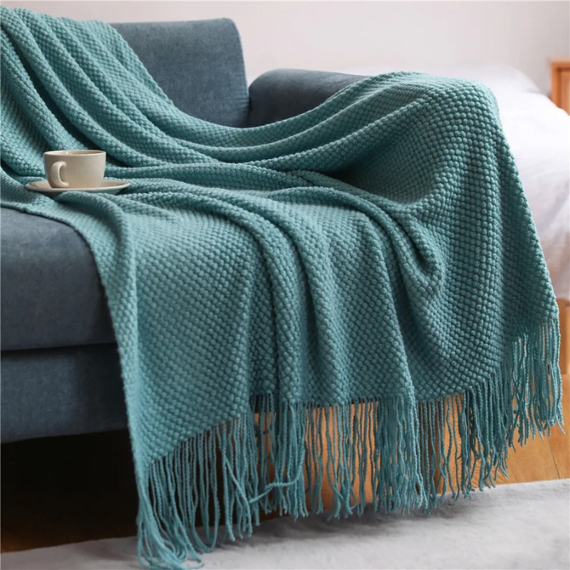 Teal knit throw blanket with long fringe tassels, draped over a couch arm