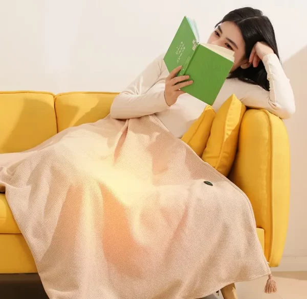 Beige heated shawl wrap with zip front and tasseled edge, laid over sofa